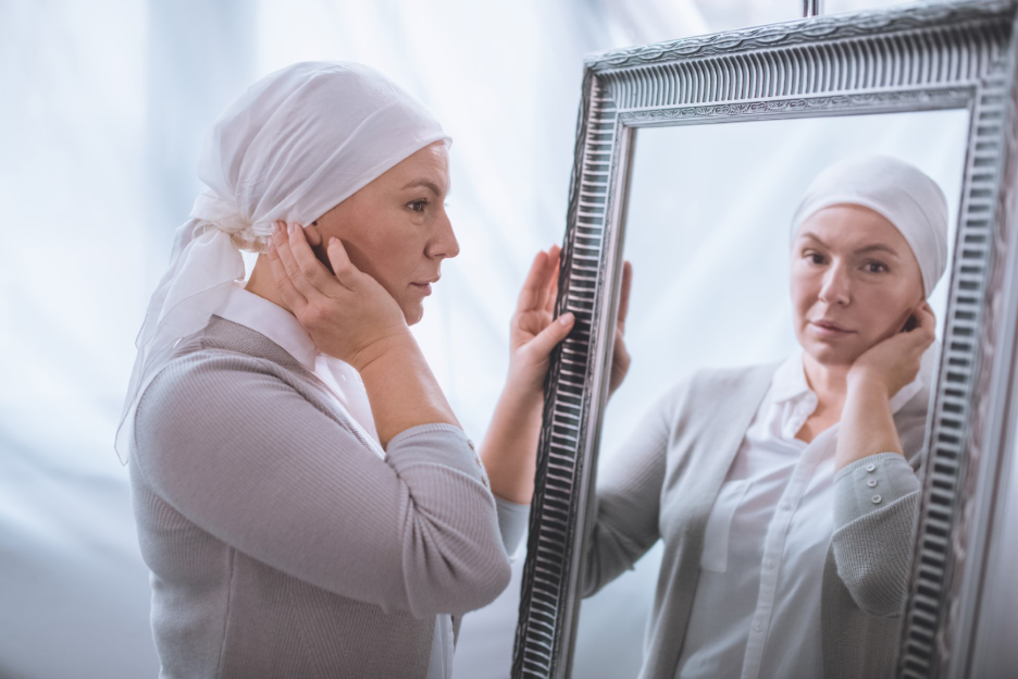 Woman undergoing cancer treatment looks at her reflection in a mirror while wearing a head covering.